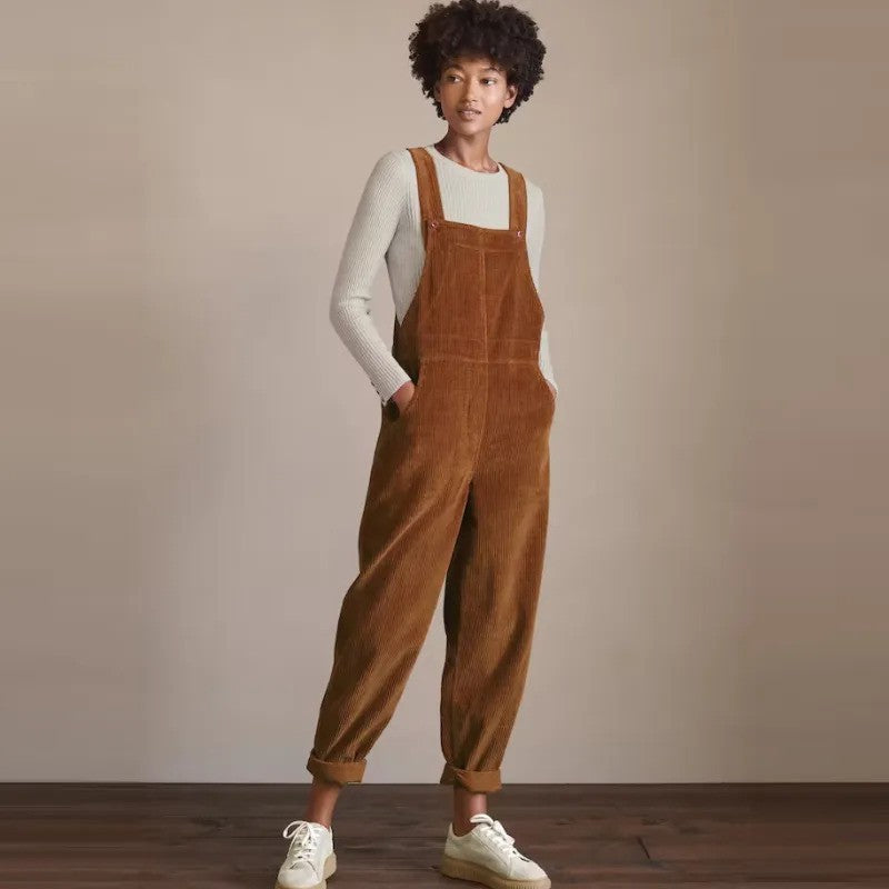 Une femme portant une salopette chaude en velours côtelé couleur rouille, associée à un haut à manches longues côtelé de couleur crème et des baskets blanches. La tenue offre un look d'inspiration vintage, alliant confort et style dans une ambiance décontractée et casual.