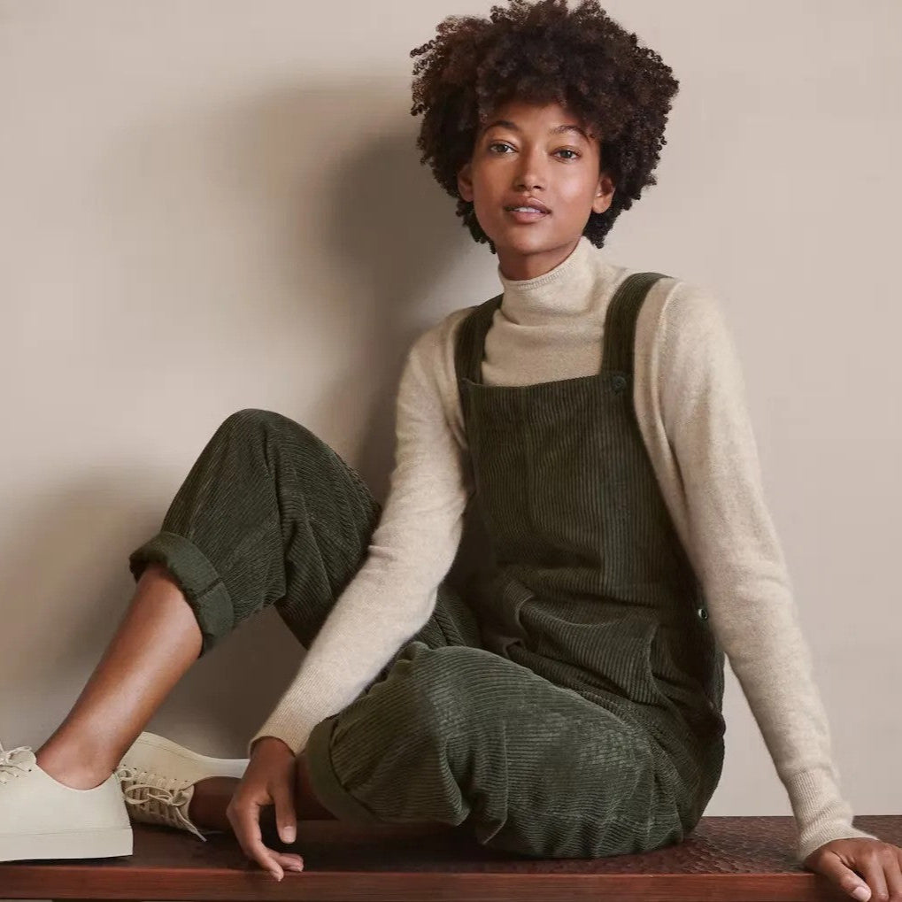 Une femme élégante portant une salopette en velours côtelé vert olive, associée à un pull à col roulé beige et des baskets blanches. Le look est à la fois décontracté et chic, évoquant une esthétique vintage, parfaite pour la mode automne-hiver.