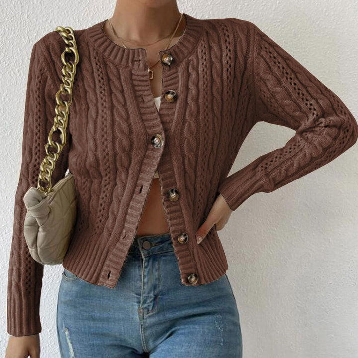 Femme portant un cardigan texturé marron à motifs torsadés, légèrement ouvert sur un haut blanc et assorti à un jean taille haute. Le modèle met en valeur une maille détaillée, des boutons décoratifs et une coupe moderne et élégante, complétée par un sac à chaîne dorée.