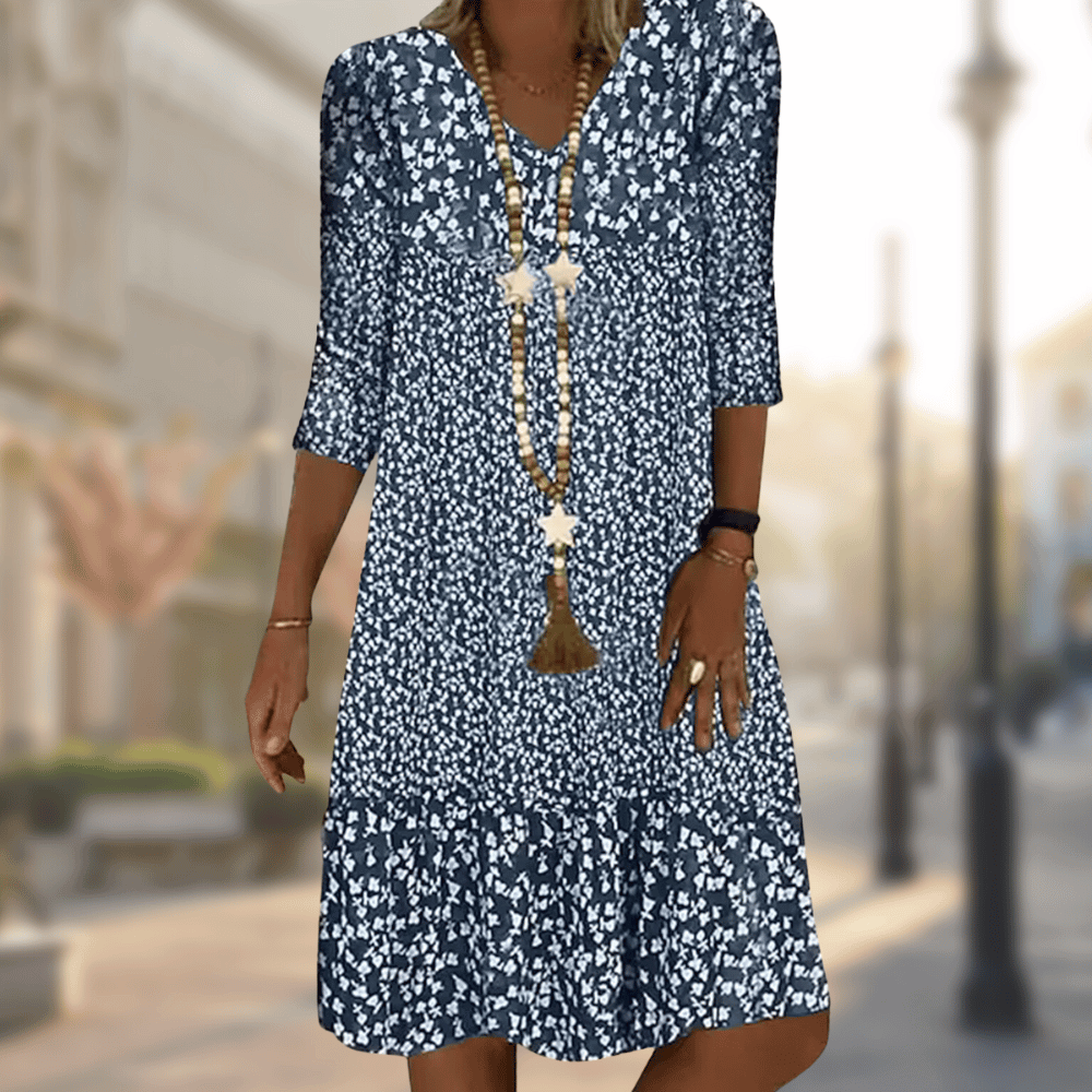 Femme portant la robe midi moderne Oriane bleu marine à imprimé floral blanc, coupe ample avec col en V, manches 3/4 et volant au bas, accessoirisée avec un long collier, en extérieur.