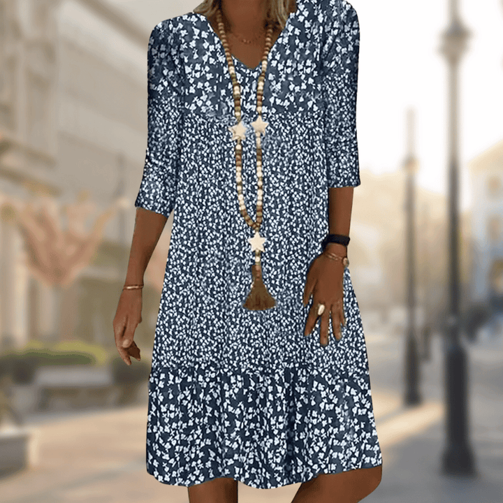 Femme portant la robe midi moderne Oriane bleu marine à imprimé floral blanc, coupe ample avec col en V, manches 3/4 et volant au bas, accessoirisée avec un long collier, en extérieur.