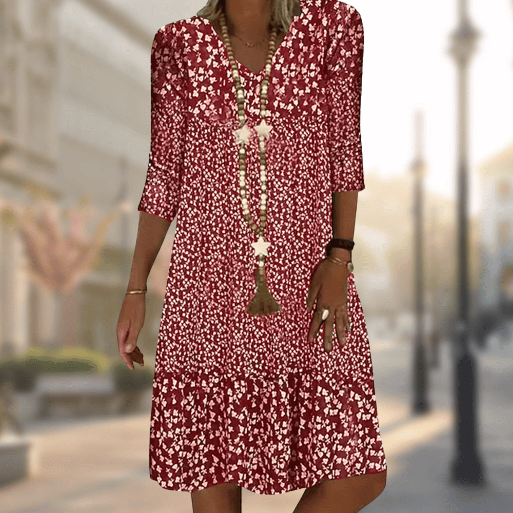 Femme portant la robe midi moderne Oriane bordeaux à imprimé floral blanc, coupe ample avec col en V, manches 3/4 et volant au bas, accessoirisée avec un long collier, en extérieur.