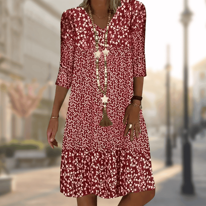 Femme portant la robe midi moderne Oriane bordeaux à imprimé floral blanc, coupe ample avec col en V, manches 3/4 et volant au bas, accessoirisée avec un long collier, en extérieur.