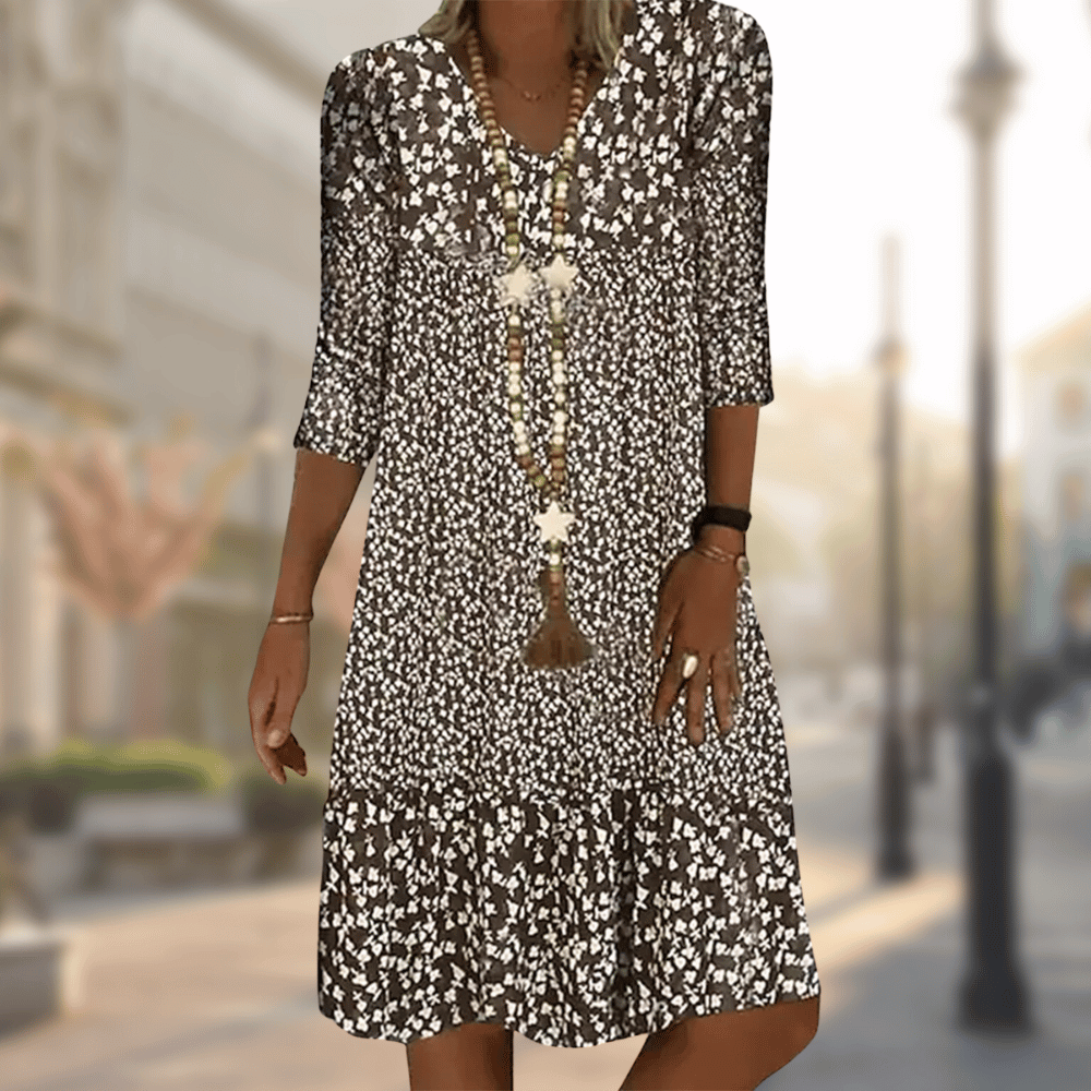 Femme portant la robe midi moderne Oriane noire à imprimé floral blanc, coupe ample avec col en V, manches 3/4 et volant au bas, accessoirisée avec un long collier, en extérieur.