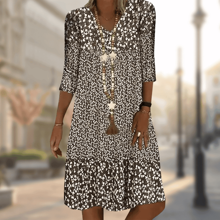 Femme portant la robe midi moderne Oriane noire à imprimé floral blanc, coupe ample avec col en V, manches 3/4 et volant au bas, accessoirisée avec un long collier, en extérieur.