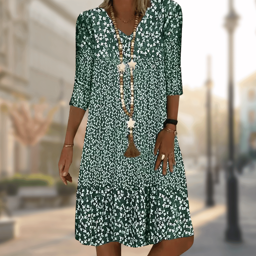 Femme portant la robe midi moderne Oriane verte à imprimé floral blanc, coupe ample avec col en V, manches 3/4 et volant au bas, accessoirisée avec un long collier, en extérieur.