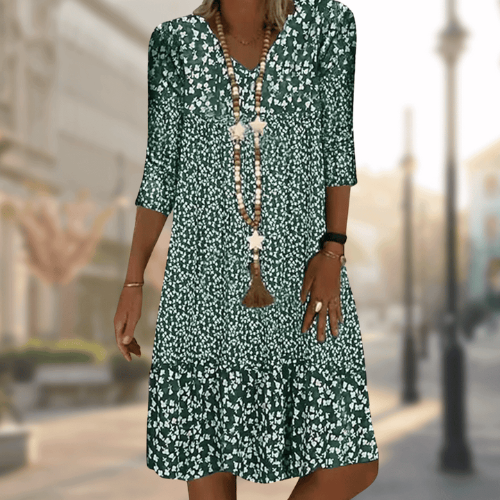 Femme portant la robe midi moderne Oriane verte à imprimé floral blanc, coupe ample avec col en V, manches 3/4 et volant au bas, accessoirisée avec un long collier, en extérieur.