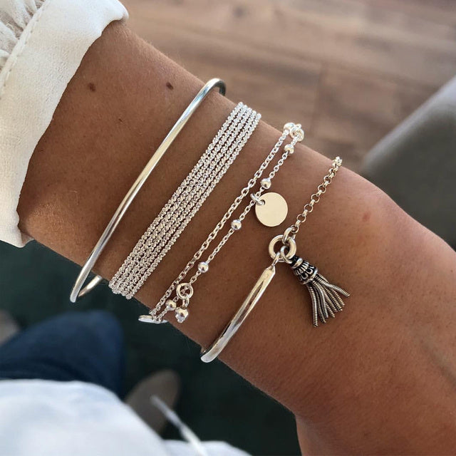 Bracelets en argent portés sur un poignet, incluant plusieurs chaînes fines, un bracelet avec un petit pendentif circulaire et un autre avec un charm à franges. Un design boho élégant et moderne.