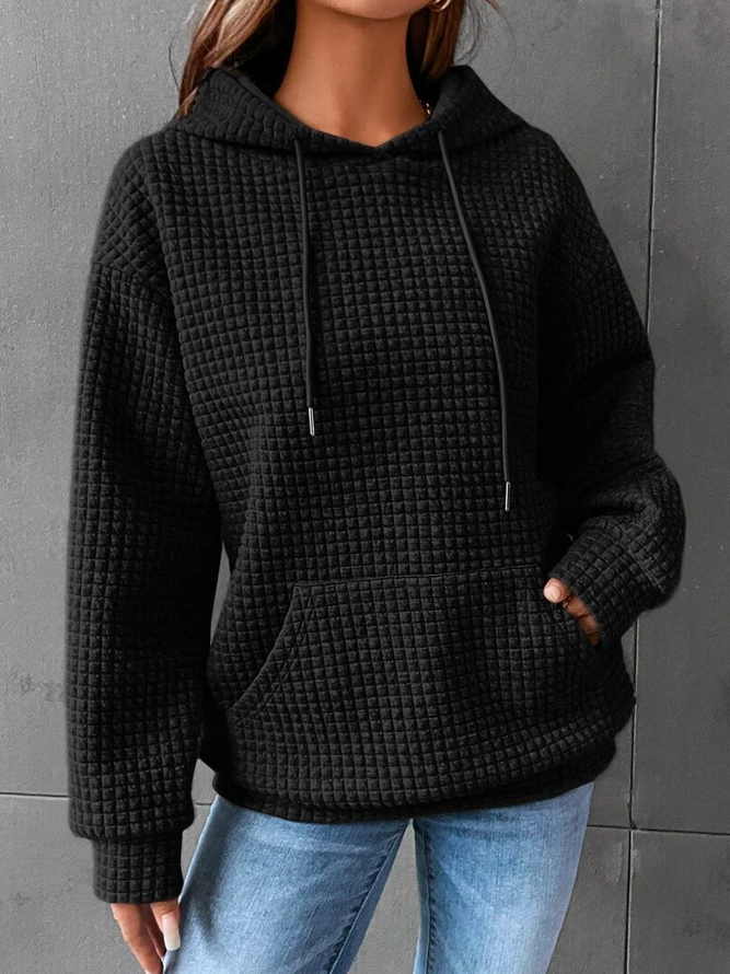 Une femme porte un sweat à capuche noir en maille gaufrée, à la coupe ample et décontractée. Ce vêtement est doté d'une grande poche kangourou sur le devant et de cordons de serrage à la capuche. Son tissu épais et structuré lui confère un style moderne et confortable, idéal pour l'hiver. Le fond gris met en valeur la texture du sweat et son allure à la fois élégante et douillette.