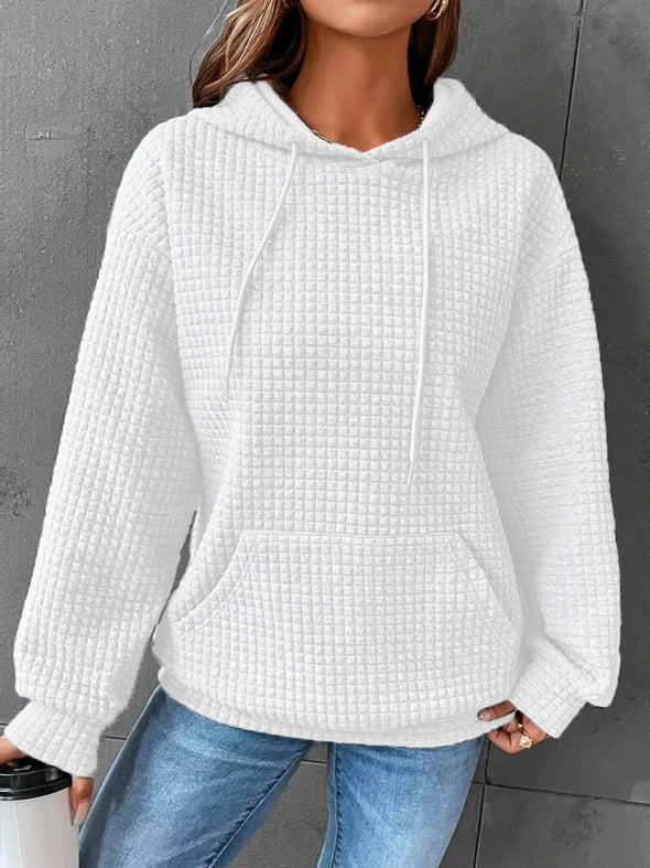 Femme portant un sweat à capuche blanc à texture gaufrée, avec une coupe ample et décontractée. Le vêtement est doté d’une grande poche kangourou à l’avant et de cordons de serrage assortis à la capuche. Le tissu épais et moelleux procure un style cosy et moderne, idéal pour les journées froides. Le fond gris met en valeur la couleur claire du sweat et la douceur de sa texture.