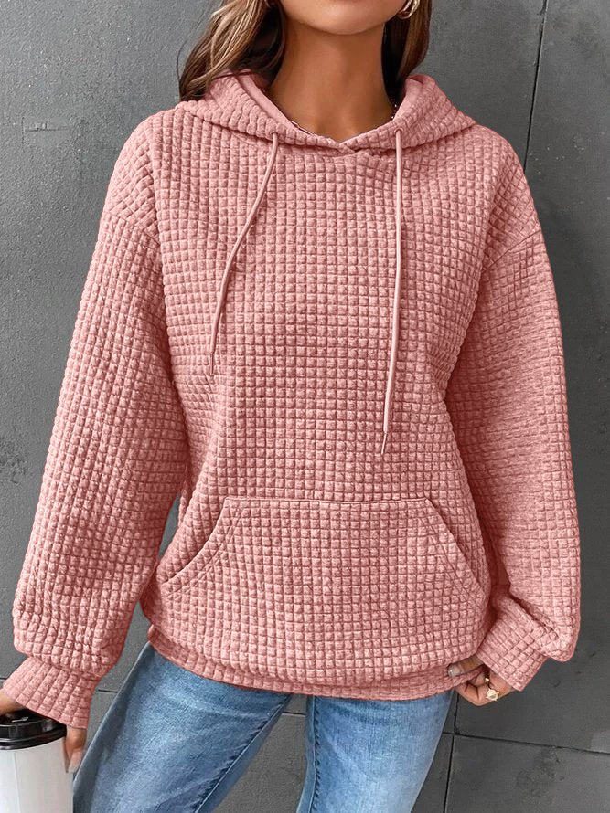Une femme porte un sweat à capuche rose à texture gaufrée, doté d'une grande poche kangourou et de cordons de serrage assortis. Sa coupe ample et décontractée est idéale pour un style moderne et confortable. Le tissu épais et doux offre chaleur et confort, parfaits pour l'hiver. Le fond gris met en valeur la teinte douce et féminine du sweat.