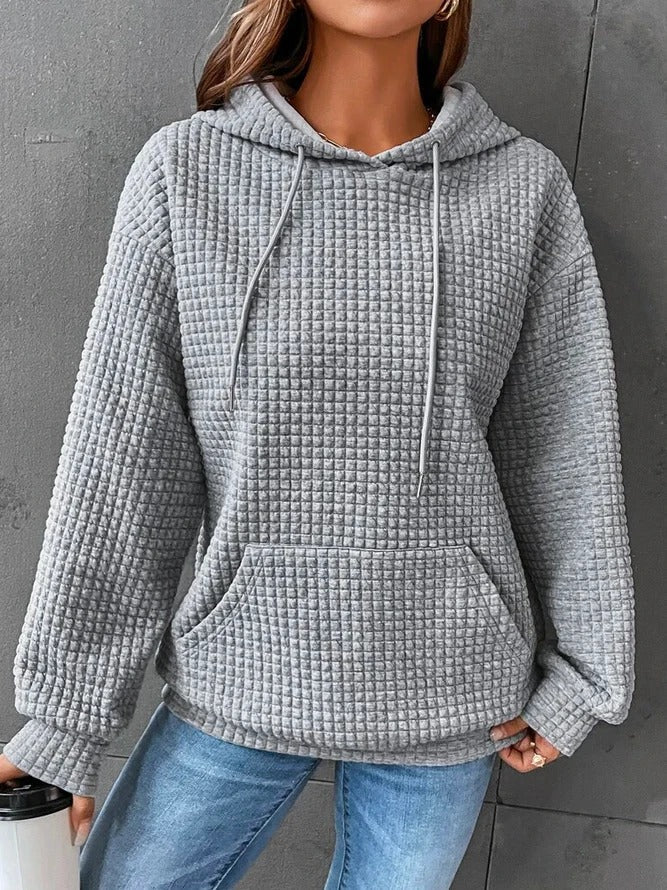 Une femme porte un sweat à capuche gris gaufré à la coupe ample et décontractée. Il est doté d'une grande poche kangourou sur le devant et de cordons de serrage assortis à la capuche. Son tissu épais et doux offre un confort idéal par temps frais. Le fond gris met en valeur la texture structurée du sweat et son style moderne et douillet.