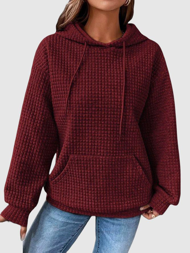Une femme porte un sweat à capuche rouge à texture gaufrée, doté d'une grande poche kangourou et de cordons de serrage assortis. Sa coupe ample et décontractée est idéale pour un style moderne et confortable. Le tissu épais et doux offre chaleur et confort, parfaits pour les journées d'hiver. Le fond clair met en valeur la couleur profonde du sweat et la texture du tissu.