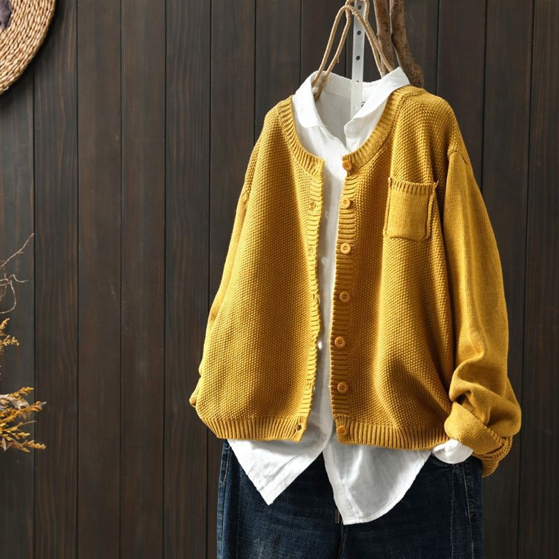 Cardigan oversize pour femme de couleur jaune moutarde, suspendu sur un cintre au-dessus d’une chemise blanche ample. Le modèle présente une maille texturée, des boutons assortis et une coupe large, offrant un style moderne, lumineux et décontracté.