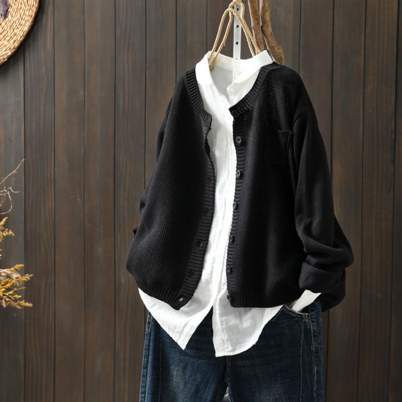 Cardigan oversize pour femme de couleur noir, suspendu sur un cintre au-dessus d’une chemise blanche ample. Le modèle présente une maille texturée, des boutons assortis et une coupe large, offrant un style moderne, minimaliste et décontracté.