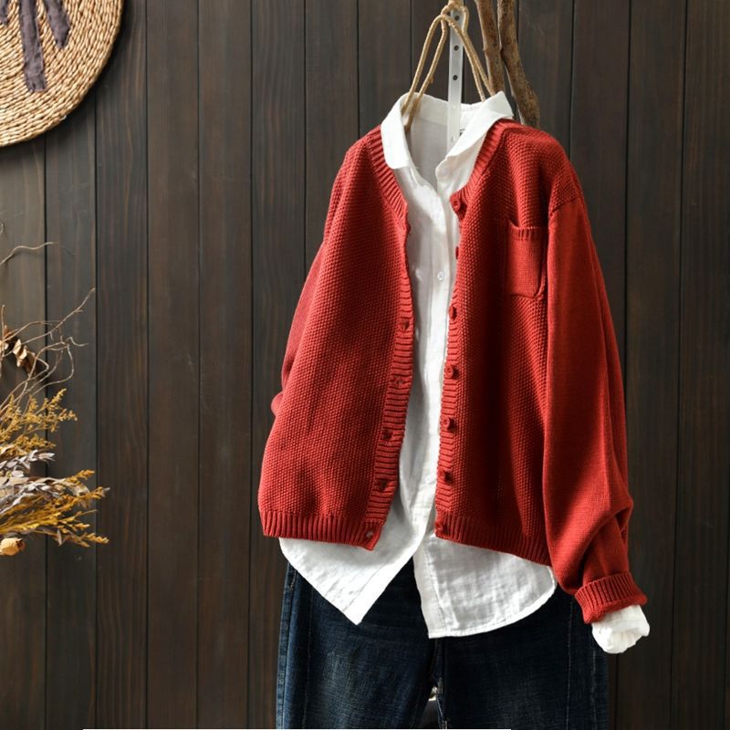 Cardigan oversize pour femme de couleur rouge brique, suspendu sur un cintre au-dessus d’une chemise blanche ample. Le modèle présente une maille texturée, des boutons assortis et une coupe large, offrant un style moderne, chaleureux et décontracté.