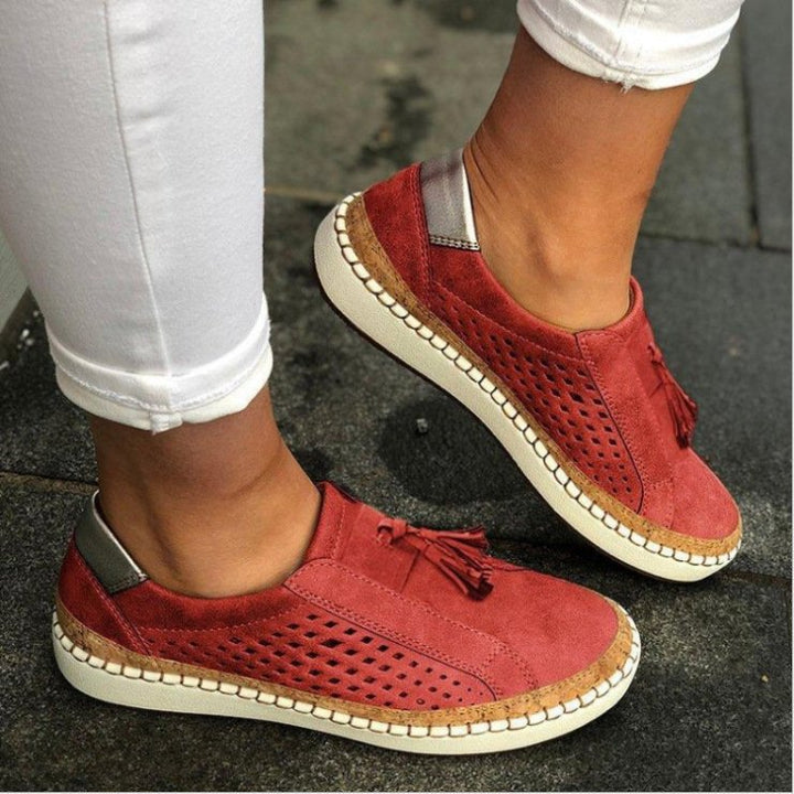Pantoufles plates rouge brique en suédine perforée avec pampilles, semelle épaisse blanche et liseré effet liège, portées avec un pantalon blanc.