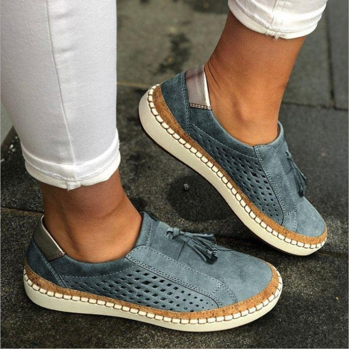 Pantoufles plates bleu gris en suédine perforée avec pampilles, semelle épaisse blanche et liseré effet liège, portées avec un pantalon blanc.