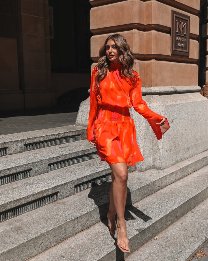 Robe mini d'été à manches longues pour femme, de style moderne avec un dégradé de couleurs orange et rose. Le modèle présente un buste smocké et des manches évasées, porté par une femme marchant dans une rue ensoleillée, créant une silhouette élégante et dynamique.
