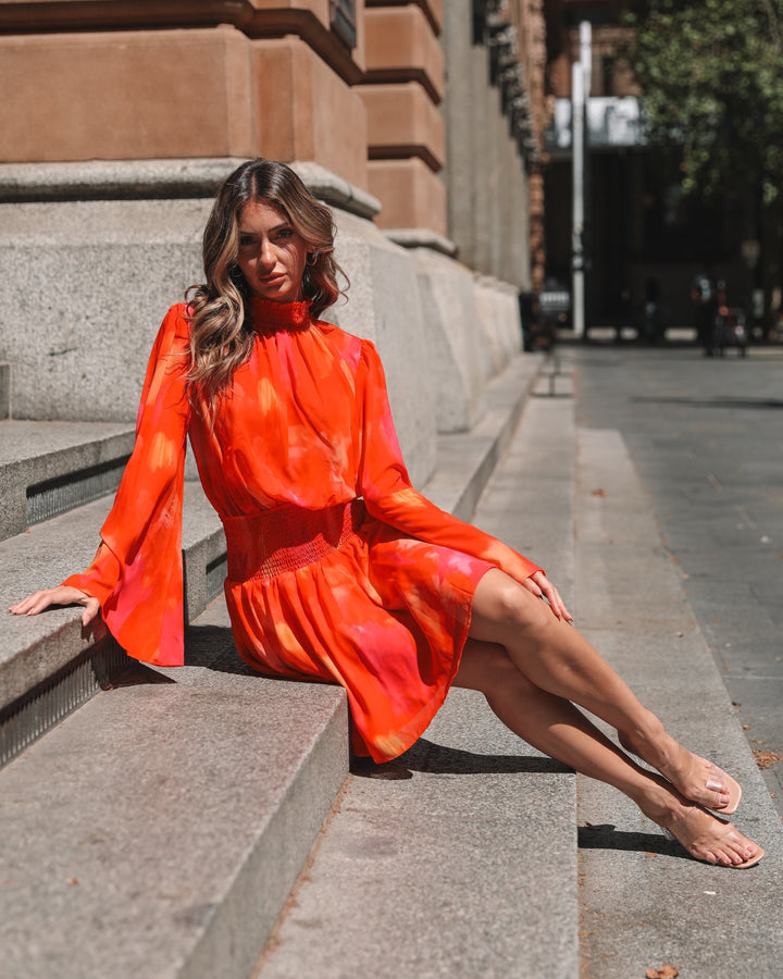 Robe mini d'été à manches longues pour femme, coloris dégradé orange et rose, avec des manches évasées et un buste smocké. Le modèle est porté par une femme assise sur des marches en pierre dans un cadre urbain, affichant un style décontracté et élégant, parfait pour les journées ensoleillées.
