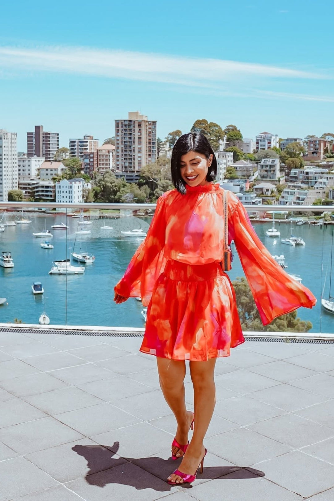 Robe mini d'été à manches longues pour femme, avec un dégradé vibrant de rose et orange. Le modèle présente un buste smocké et des manches évasées, portée par une femme souriante, se tenant sur une terrasse avec une vue sur le port et les bâtiments environnants. Parfaite pour un look estival chic et dynamique.