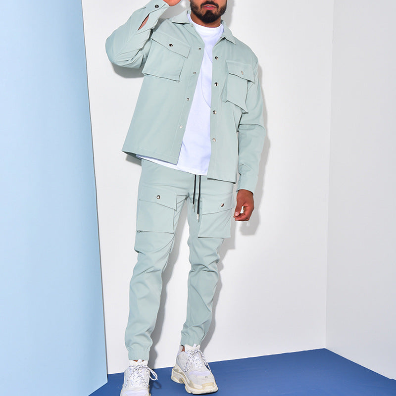 Homme portant un ensemble cargo vert pâle avec une veste à poches et un pantalon assorti. Il ajuste sa montre tout en se tenant devant un fond blanc et bleu. Il porte également un t-shirt blanc et des baskets blanches.