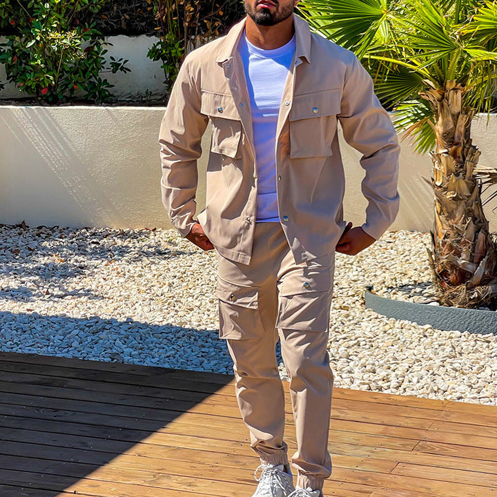 Homme marchant en extérieur, portant un ensemble cargo de couleur beige, composé d’une veste à poches et d’un pantalon assorti. Il porte également un t-shirt blanc et des baskets blanches, déambulant sur un sol en bois, avec un fond naturel composé de palmiers et de pierres décoratives.