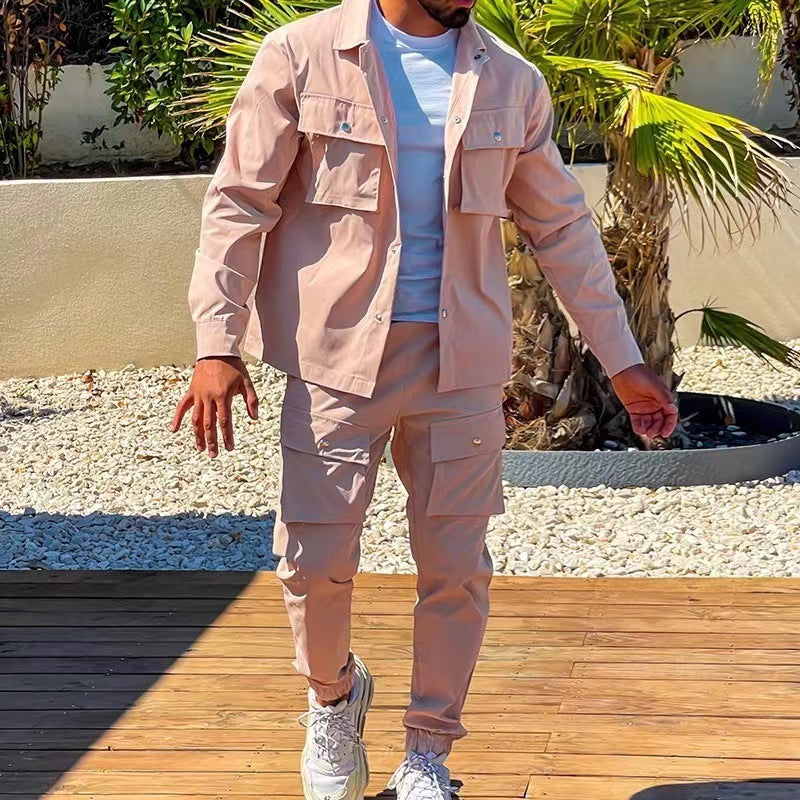 Homme portant un ensemble cargo élégant de couleur beige clair, composé d’une veste à poches et d’un pantalon assorti. Il est debout à l’extérieur sur une terrasse en bois, près d’une piscine et entouré de palmiers. Il porte un t-shirt blanc et des baskets modernes, créant un look décontracté et raffiné parfait pour les journées ensoleillées.