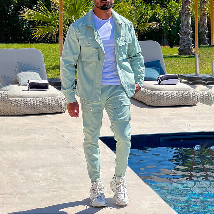 Un homme se tient près d’une piscine, vêtu d’un ensemble cargo moderne de couleur vert clair. Sa tenue comprend une veste légère à poches multiples et un pantalon assorti, accompagnés d’un t-shirt blanc et de baskets blanches. Le look combine élégance décontractée et confort, parfait pour un style estival ou urbain. L’arrière-plan met en valeur une ambiance extérieure raffinée avec des chaises longues et des palmiers.