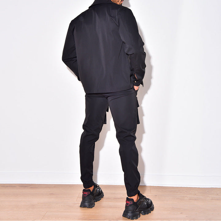 Homme portant un ensemble sportif et élégant de couleur noire, composé d'une veste et d'un pantalon cargo. Il est vu de dos dans un cadre épuré, avec un sol en bois clair, mettant en avant le style moderne et fonctionnel du vêtement. L'ensemble est complété par des baskets noires, apportant une touche dynamique au look.