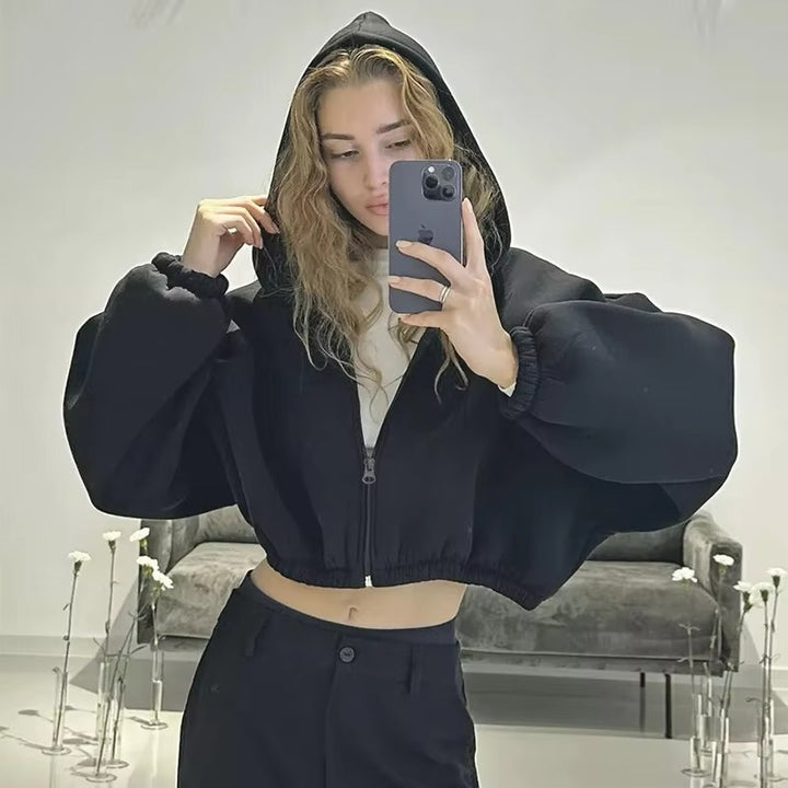 Femme portant un sweat à capuche court noir avec fermeture éclair, manches larges et ourlet élastique, prenant un selfie dans un intérieur moderne. Elle tient la capuche d’une main et son téléphone de l’autre, dévoilant un style décontracté et tendance dans un décor minimaliste avec des fleurs blanches en arrière-plan.