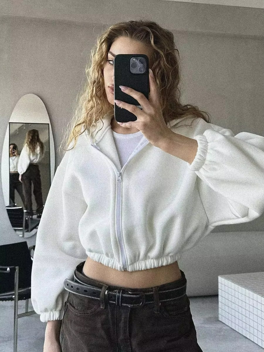 Femme portant un sweat à capuche court blanc avec fermeture éclair, manches bouffantes et taille élastique, prenant un selfie dans une pièce moderne. Son reflet apparaît dans un miroir derrière elle, et elle associe la tenue à un pantalon foncé avec une ceinture noire. L’ambiance de la photo est minimaliste et élégante.