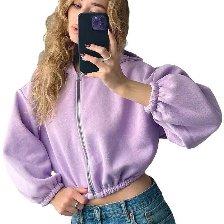 Une femme vêtue d'un sweat-shirt à capuche court violet zippé, à manches bouffantes et taille élastiquée, prend un selfie. Cheveux ondulés et jean taille basse, elle affiche un style jeune, tendance et décontracté sur fond blanc.