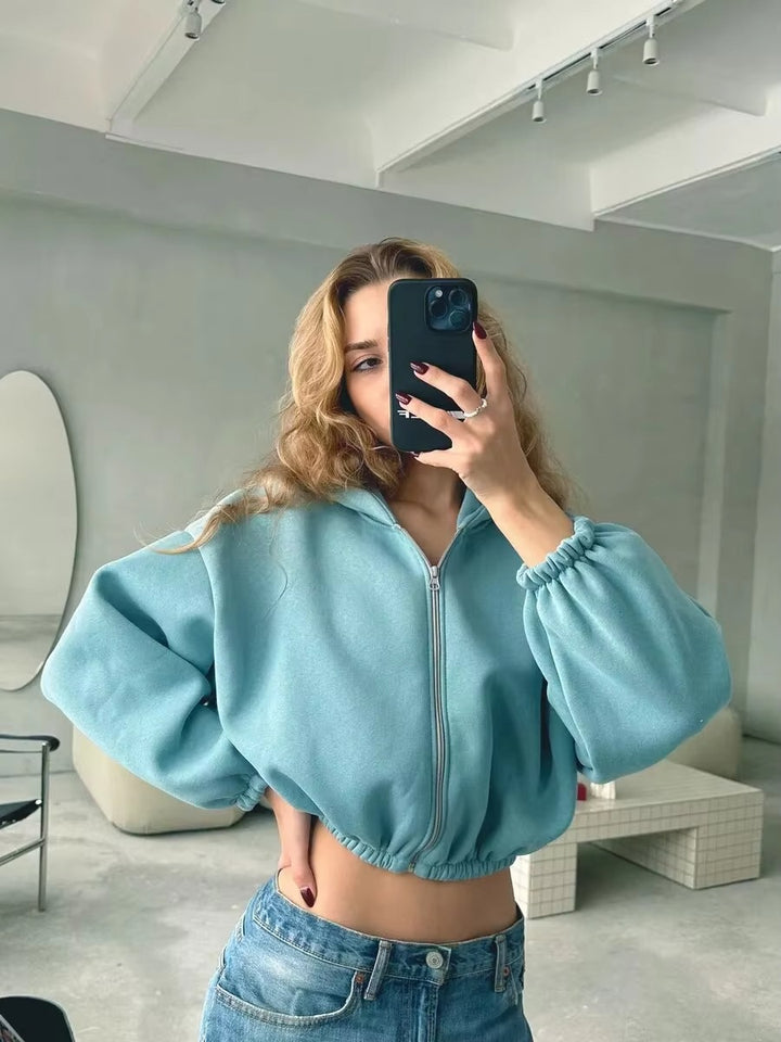 Une femme vêtue d'un sweat-shirt à capuche court bleu zippé, à manches bouffantes et taille élastiquée, prend un selfie dans une pièce moderne. Ses cheveux ondulés et son jean bleu reflètent un style décontracté et tendance dans un décor minimaliste.