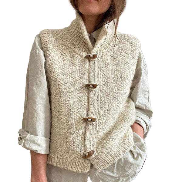 Une femme porte un gilet blanc en grosse maille, fermé par quatre boutons en bois, par-dessus une chemise ample à manches retroussées. Le vêtement présente une texture de maille travaillée et un col montant pour un style à la fois moderne et confortable.