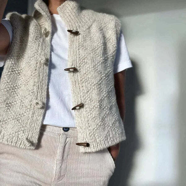 Une femme porte un gilet blanc en maille texturée, ouvert sur un t-shirt blanc et associé à un pantalon clair. Le gilet, à la coupe confortable, possède un col montant et des boutons en bois, soulignant un style moderne et décontracté.