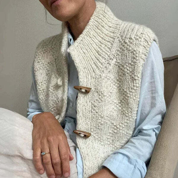 Une femme est assise, vêtue d'un gilet blanc en maille texturée, à boutons de bois et col montant, porté sur une chemise claire. La texture de la maille et la coupe souple créent un style à la fois chaleureux et élégant.