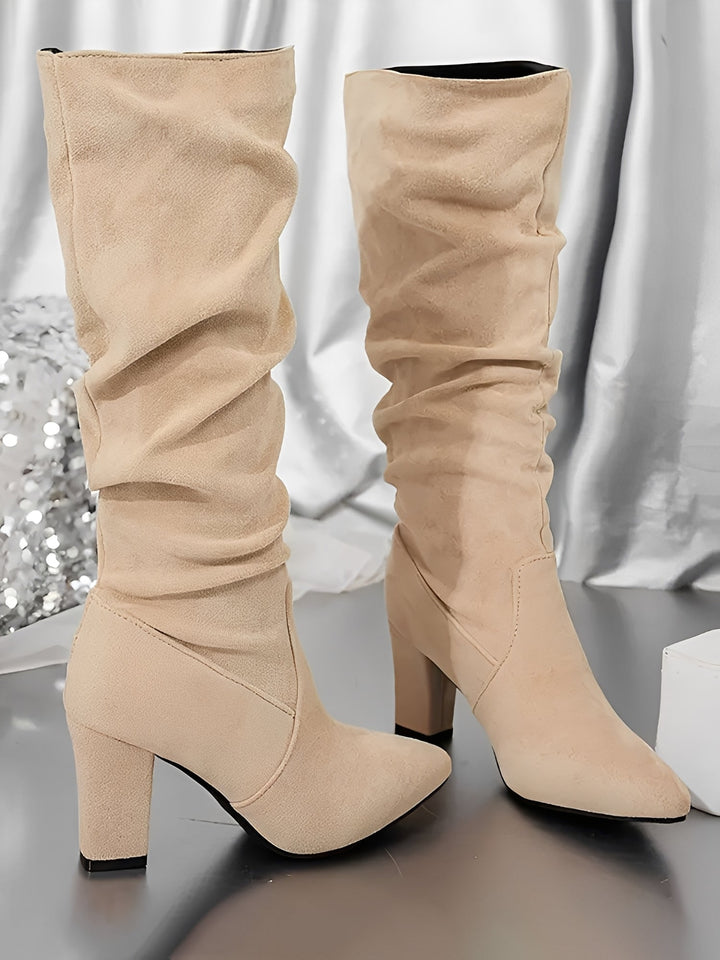 Paire de bottes hautes en daim beige posées sur une surface brillante, avec un talon épais et une tige plissée. Les bottes présentent une silhouette élégante et moderne, mises en valeur dans un décor intérieur lumineux aux accents métallisés.