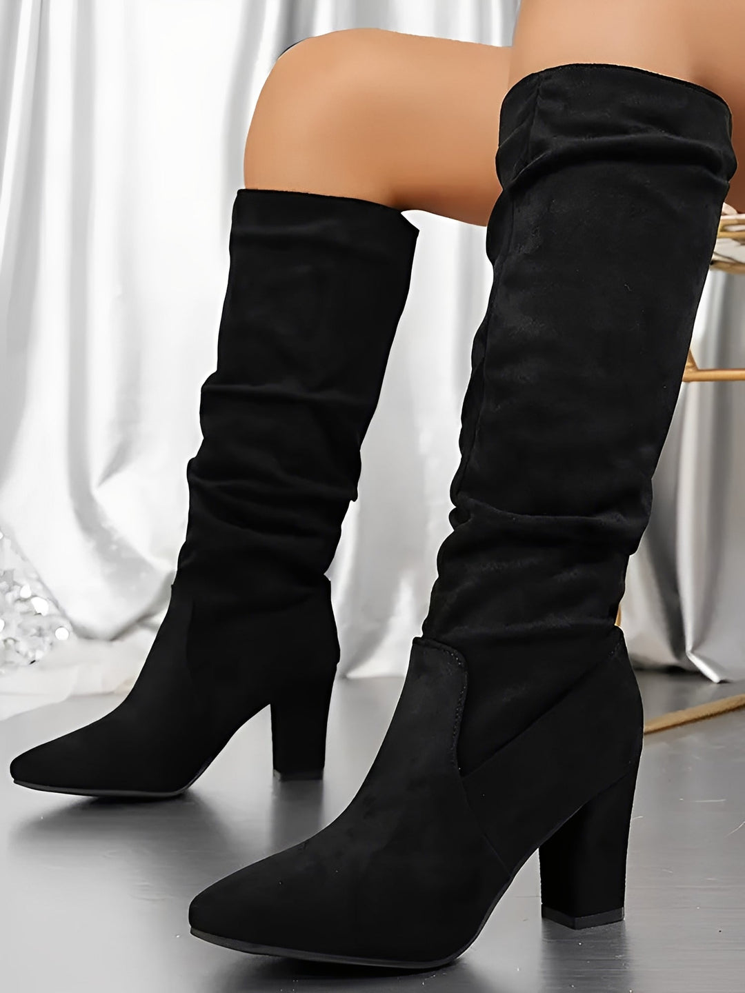 Bottes hautes en daim noir portées par une femme assise, montrées en gros plan dans un décor intérieur élégant. Les bottes présentent un talon épais, une tige légèrement plissée et une silhouette moderne, offrant un style chic et contemporain.
