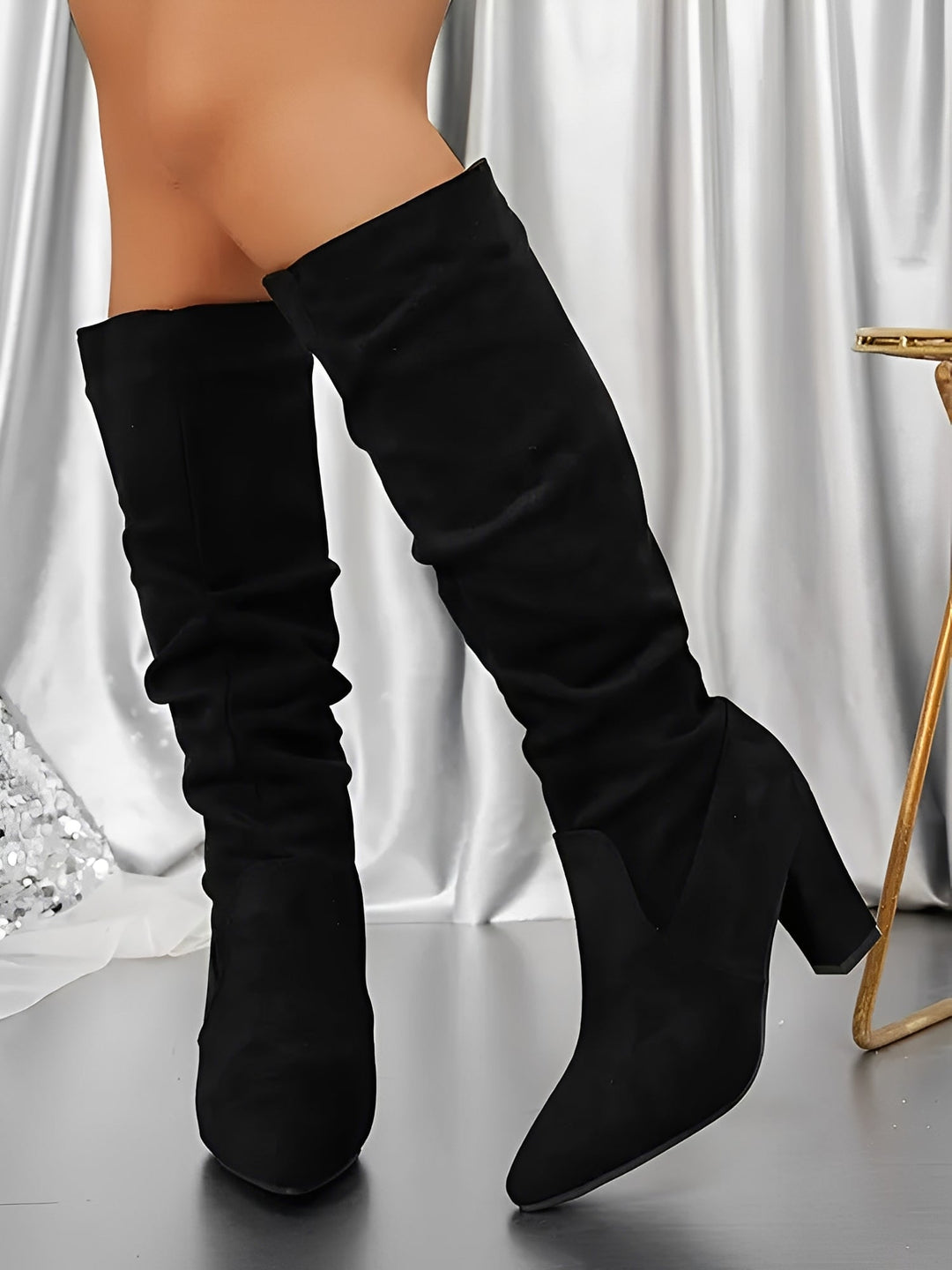 Bottes hautes en daim noir portées par une femme debout, présentées en gros plan dans un décor intérieur élégant. Le modèle possède un talon épais, une tige lisse légèrement plissée et une silhouette moderne, offrant un style chic et contemporain.