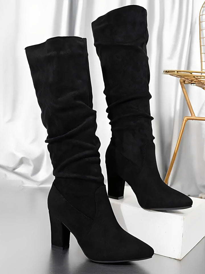 Paire de bottes hautes en daim noir présentées sur une surface lumineuse, avec un talon épais élégant et une tige légèrement plissée. Les bottes affichent une silhouette moderne et raffinée, mises en valeur dans un décor intérieur aux tons métallisés.