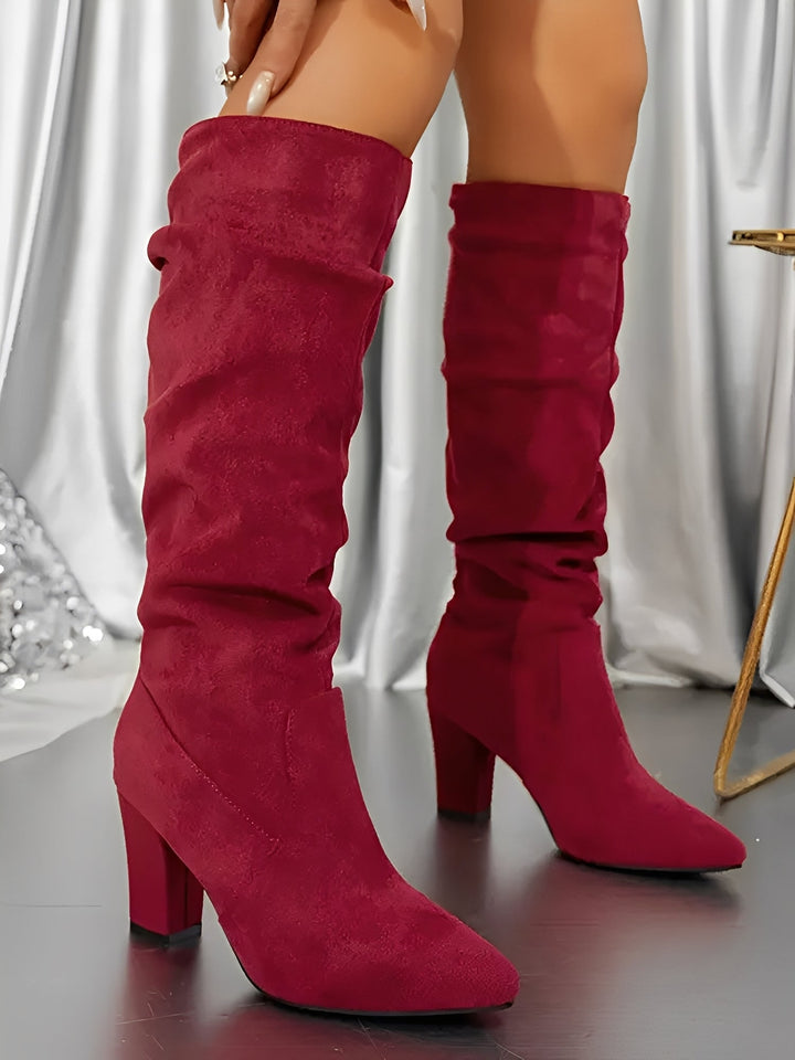 Bottes hautes en daim rouge portées par une femme debout, présentées en gros plan dans un décor intérieur élégant. Le modèle comporte un talon épais, une tige légèrement plissée et une finition moderne qui apporte une touche chic et audacieuse.