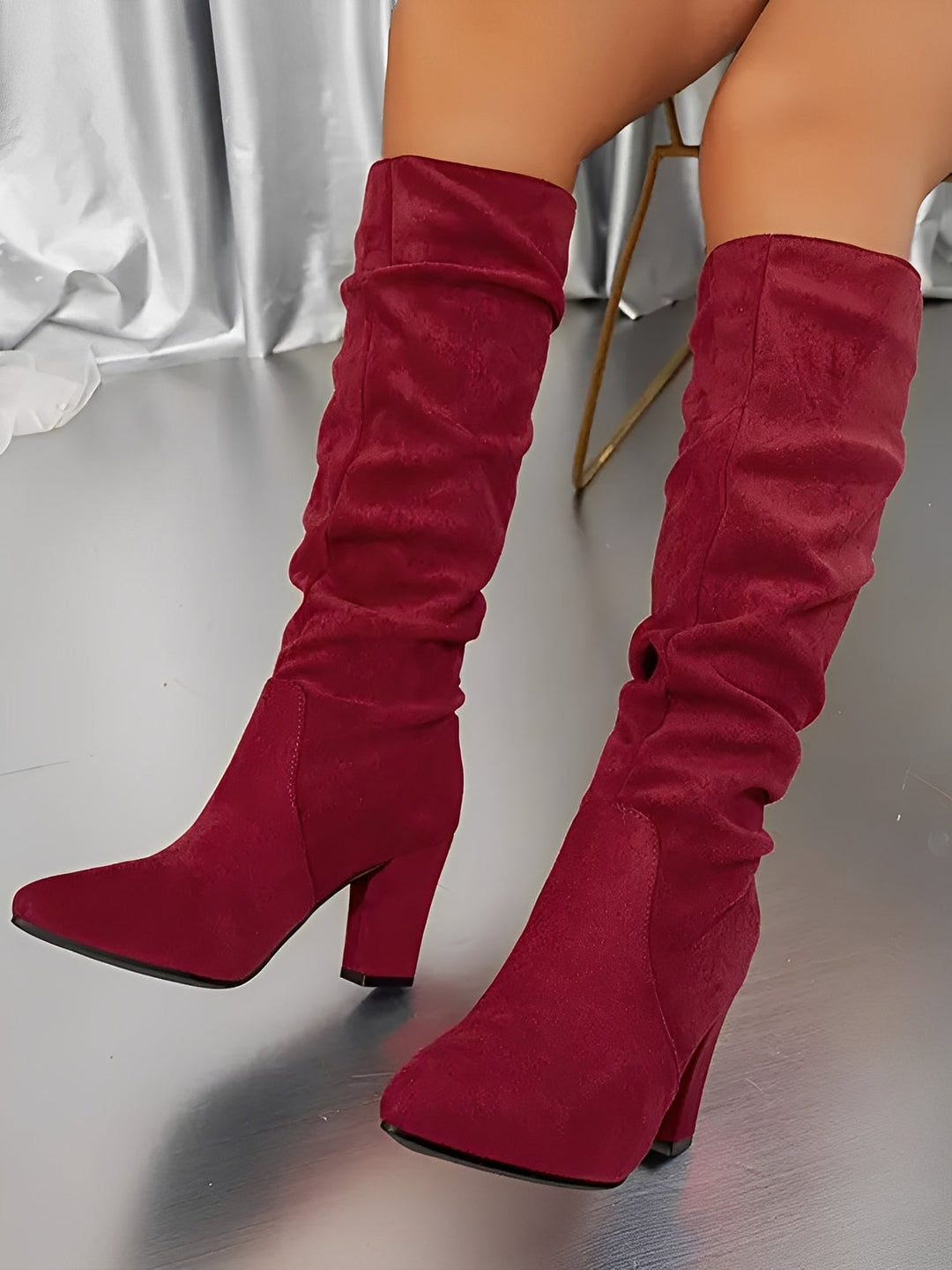 Bottes hautes en daim rouge portées par une femme assise, montrées en gros plan dans un décor intérieur moderne. Les bottes possèdent un talon épais, une tige plissée et une silhouette élégante, apportant une touche audacieuse et contemporaine.
