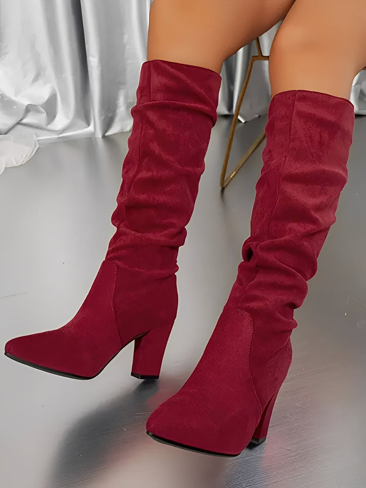 Bottes hautes en daim rouge portées par une femme assise, montrées en gros plan dans un décor intérieur moderne. Les bottes possèdent un talon épais, une tige plissée et une silhouette élégante, apportant une touche audacieuse et contemporaine.