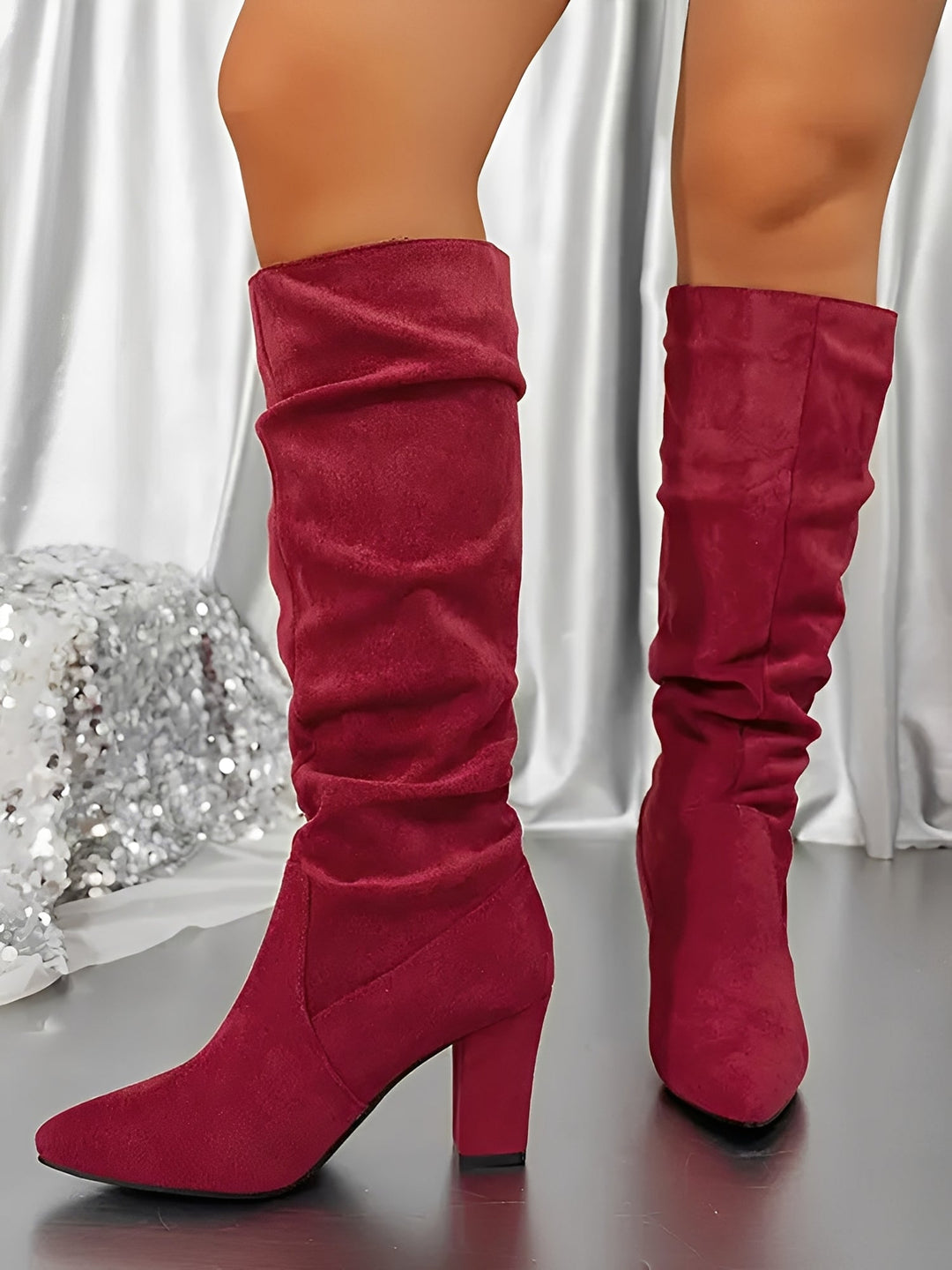 Bottes hautes en daim rouge portées par une femme, montrées en gros plan dans un décor intérieur lumineux. Le modèle présente un talon épais, une tige plissée et une silhouette moderne, apportant une touche élégante et audacieuse.