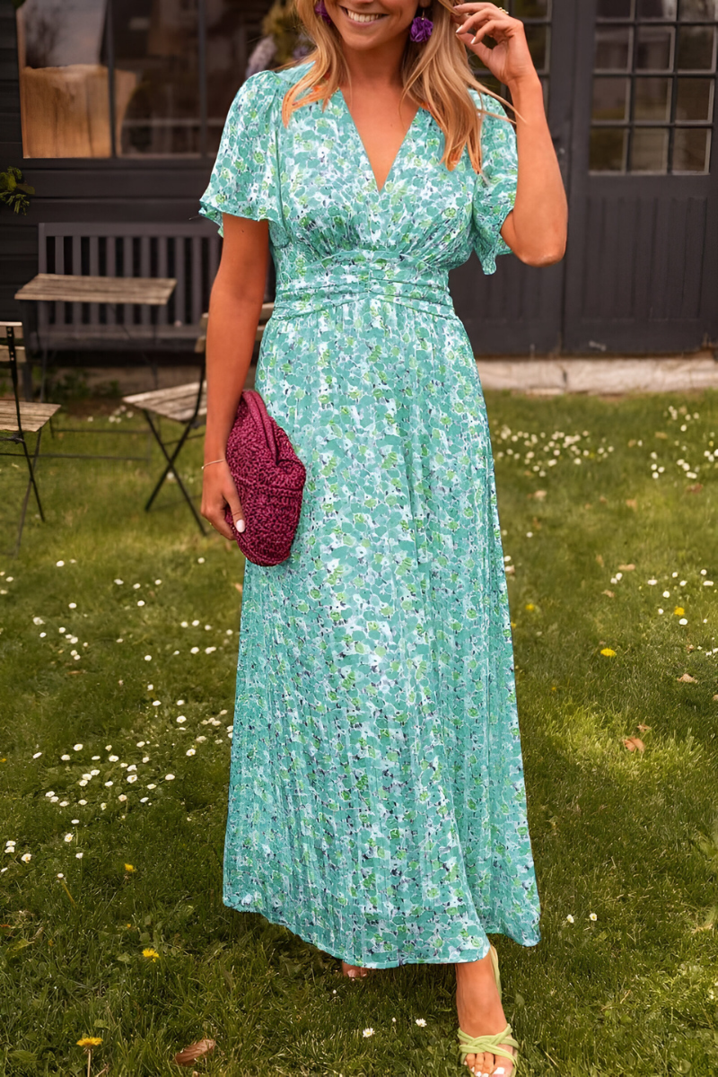 Robe longue à col en V pour femme, modèle tendance. Cette robe fluide et légère présente un joli motif floral dans des tons de vert et bleu, idéale pour les journées ensoleillées. Elle est associée à un sac en crochet violet et des sandales vertes, créant un look frais, élégant et décontracté.