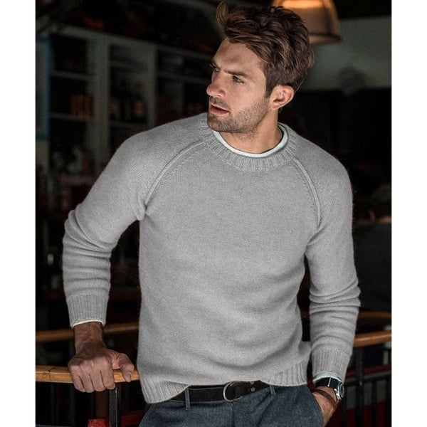 Un homme portant un pull oversize à manches longues avec des manches raglan, assorti de pantalons et d'une ceinture, se tenant dans un cadre intérieur confortable avec une expression pensive.