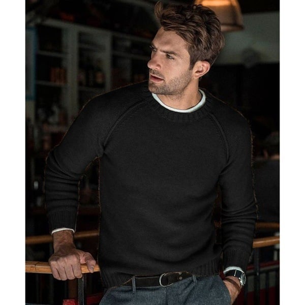 Un homme portant un pull long oversize à manches raglan, associé à un pantalon et une ceinture, se tenant dans un cadre intérieur confortable avec une expression pensante.