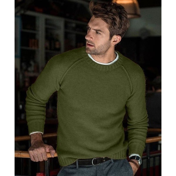 Un homme portant un pull long oversize à manches raglan, associé à un pantalon et une ceinture, se tenant avec une expression pensante dans un cadre intérieur confortable.