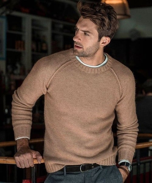 Un homme portant un pull long oversize à manches raglan, associé à un pantalon et une ceinture, se tenant avec une expression pensante.